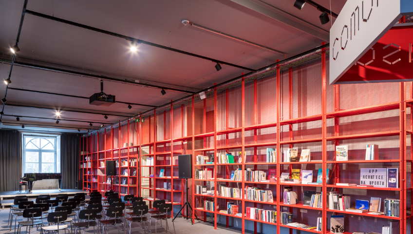 imagem da Casa Comum, um espaço com cadeiras enfileiradas ao centro e uma grande estande vermelha de libros na parede